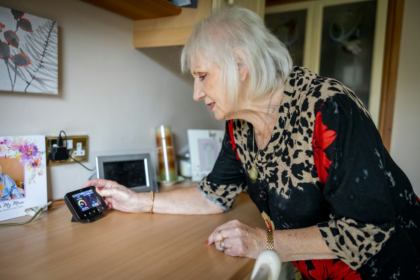 An older woman at home, attending to a small device on the counter beside a Mother's Day card